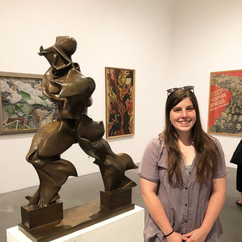 A woman with long dark brown hair and wearing a grey button up short-sleeved shirt stands in an art gallery. She has sunglasses on her head. She is Caucasian.  She stands, clasping her hands in front of her, next to a bronze futurist statue. It is 