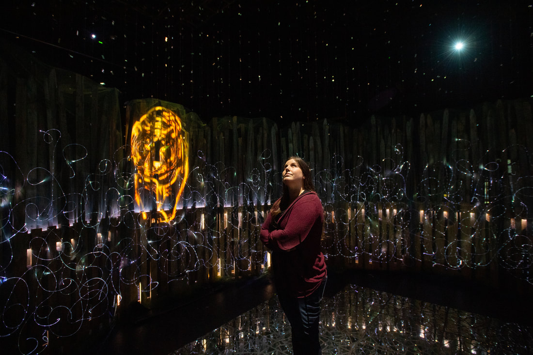 A woman with long dark hair and a crimson sweater stands on a mirror mosaic floor looking up at curtains of confetti that fill the air above a nine foot tall wooden structure built from pallets and fencing. Behind her on the wooden pallets there are swirling patterns of points of white light, created by woven fiber optics. A glowing orange tiger is projected onto the round wooden surface.